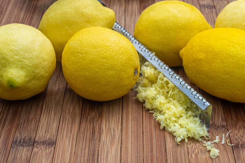 Close-up of a hand using a stainless steel zester on a lemon, with citrus zest curling off the blade